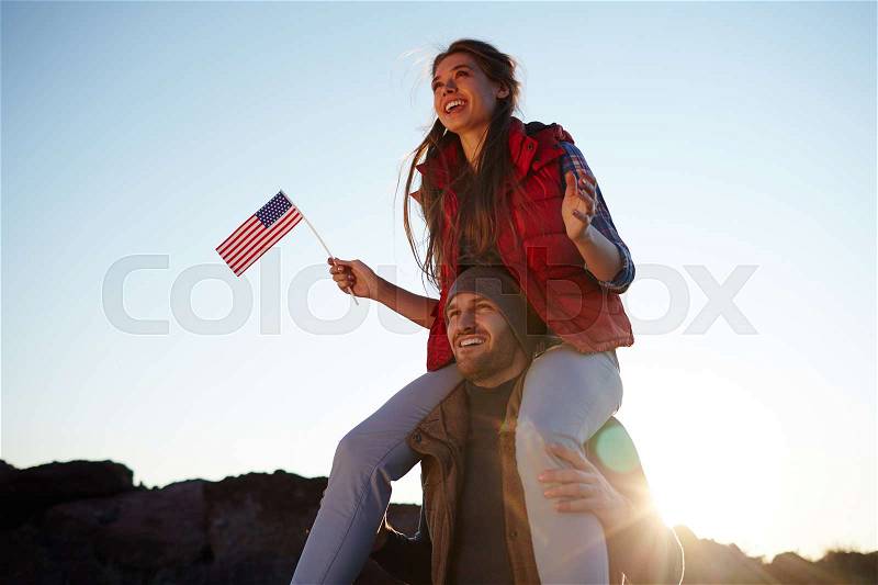 Sunlit portrait f young tourist couple celebrating American freedom: beautiful girl in hiking outwear sitting on her boyfriends shoulders smiling cheerfully and waving small American flag, stock photo