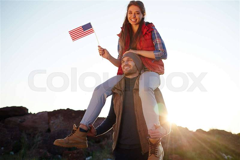 Portrait of young tourist couple celebrating American freedom: beautiful girl in hiking outwear sitting on her boyfriends shoulders smiling cheerfully to camera and waving small American flag, stock photo