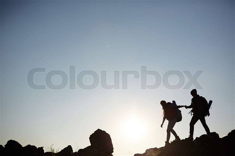 Backlit dark silhouettes of couple hiking in mountains, young man following girlfriends holding hands, side view graphic image, stock photo