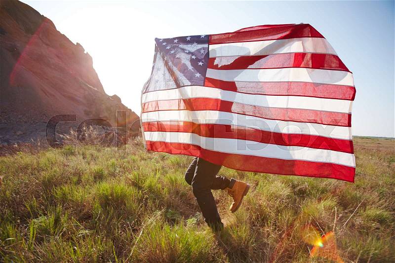 Portrait of young unrecognizable man joyfully running on green grass of mountains waving big USA banner in bright sunlight, stock photo