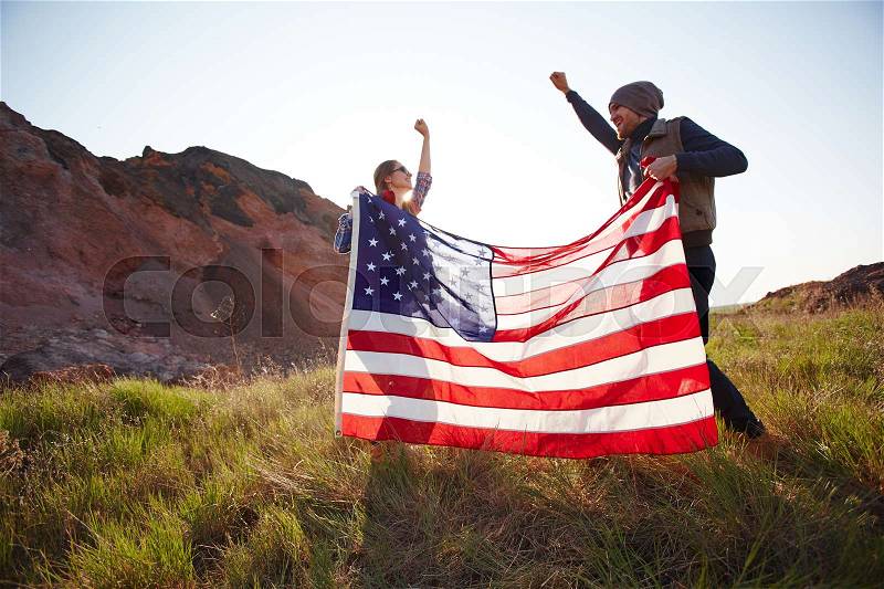 Couple of joyous young people waving big American banner jumping on top of hill in mountains, stock photo