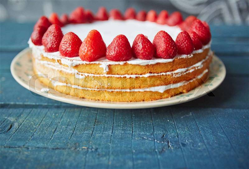 Closeup of delicious simple biscuit cake decorated with fresh ripe strawberries on top and white icing between layers standing on rustic wooden table, stock photo