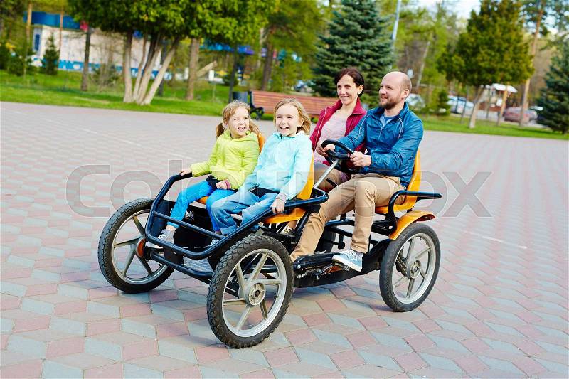 Pretty family having fun in spring park: bearded middle-aged father driving orange quadricycle pedal car while his joyful family sitting in it with wide smiles, stock photo