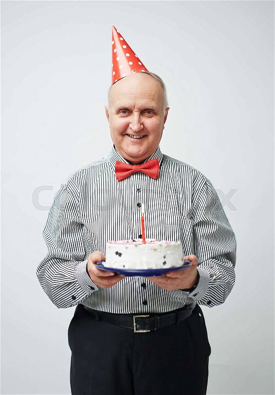 Portrait of charming old man in party hat and bow tie standing straight, holding birthday cake with one candle and smiling cheerfully, stock photo