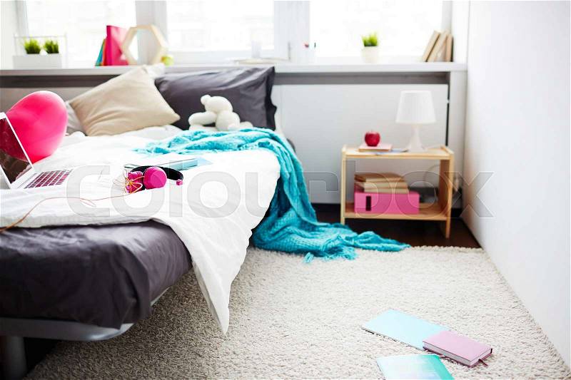 Modern messy bedroom of teenage girl: comfortable bed with laptop, headphones, blanket hanging down and school books lying on floor, stock photo