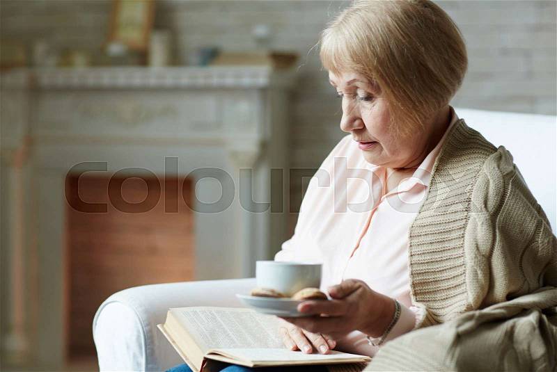 Perfect winter evening: senior woman sitting by fireplace, drinking cup of tea with delicious cookies and reading interesting novel, stock photo