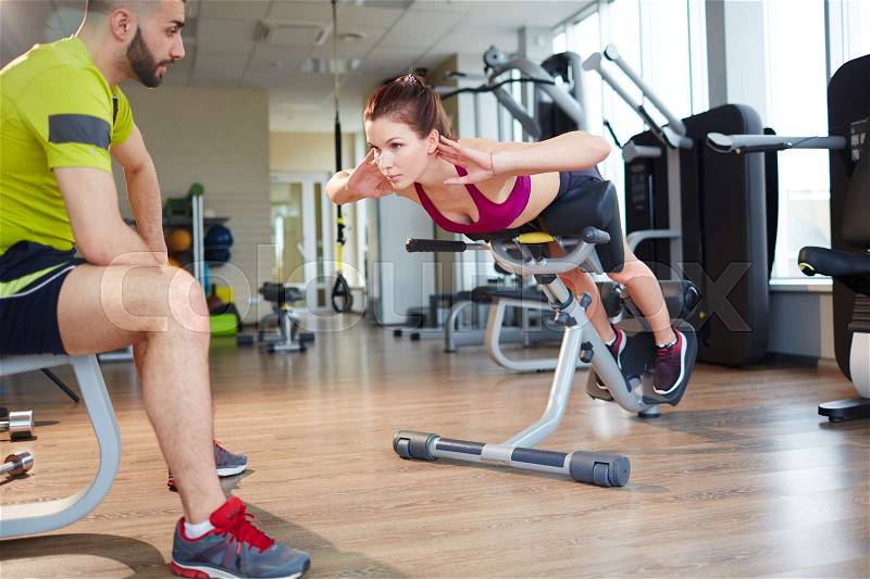 Working out in gym: handsome instructor watching young girl while performing hyperextension exercise using roman chair, stock photo