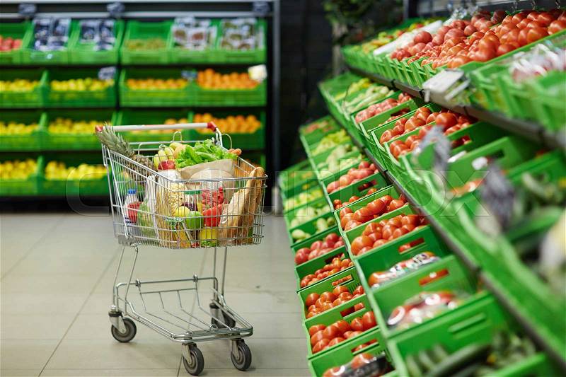 Metal shopping trolley with healthy grocery items standing in fruit and vegetable department of hypermarket, stock photo