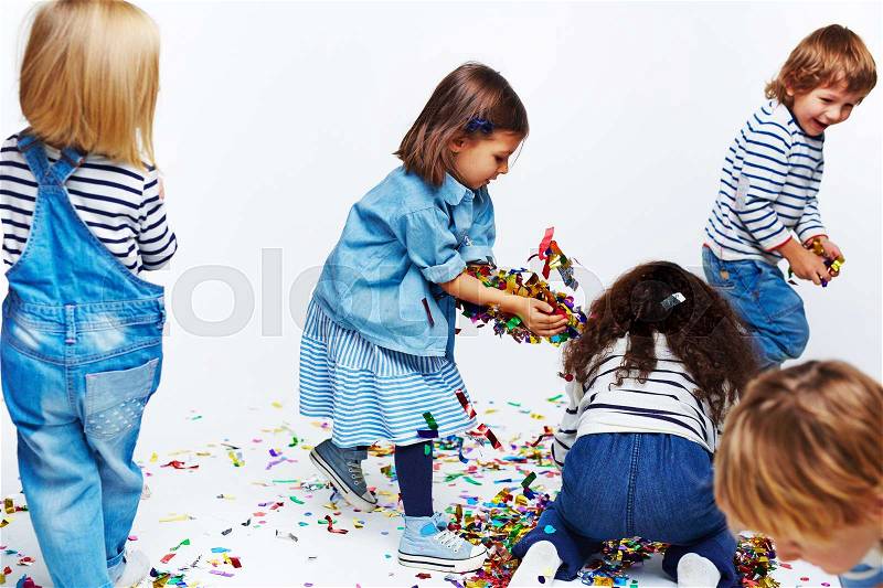 Studio shot of five various happy little kids in jeans clothes running around chaotically playing with confetti and gathering it in piles from the floor against white background, stock photo