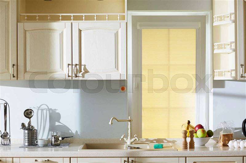 Interior of lovely beige kitchen in classic style: sink with unusual tap, wooden cupboards hanging on walls, cooking utensils and products lying on stone countertop, stock photo