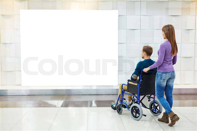 Rear view of young woman standing beside her boyfriend in wheelchair and looking at blank advertising board in mall, stock photo
