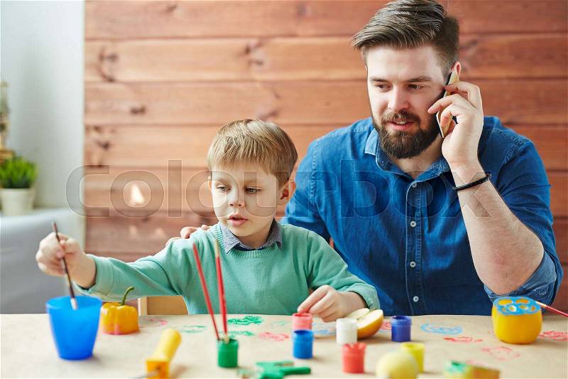 Waist-up portrait of busy bearded man talking on his mobile phone while his cute little son sitting next to him and drawing picture with gouache, stock photo