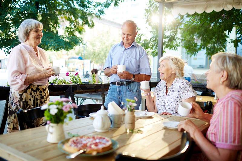 Cheerful senior friends celebrating holiday in outdoor cafe while one of them making toast; teapot and tasty pie standing on wooden table, stock photo