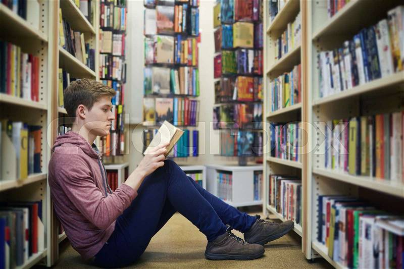 Serious guy sitting on the floor between bookshelves and reading in library, stock photo