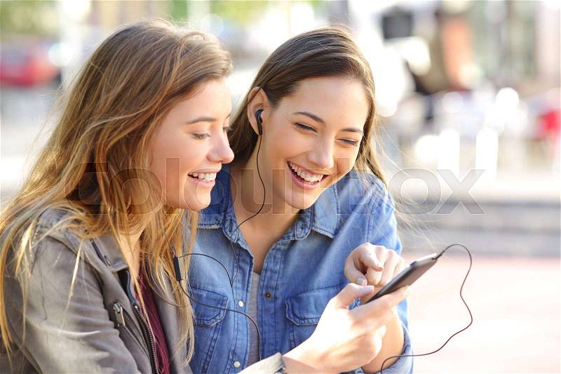 Two happy friends sharing a phone to listen music on line together in the street with an urban background, stock photo