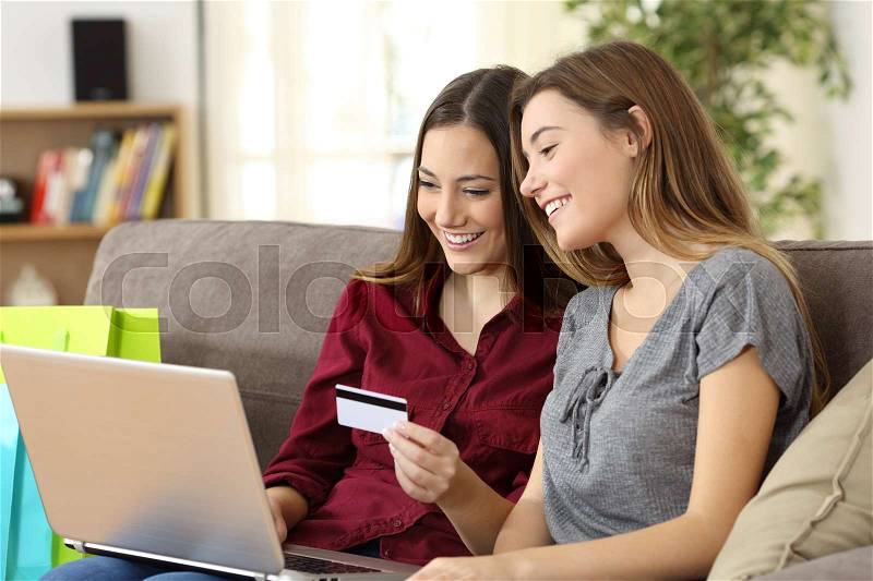 Two happy roommates buying together on line with credit card and a laptop sitting on a sofa at home with a homey background, stock photo