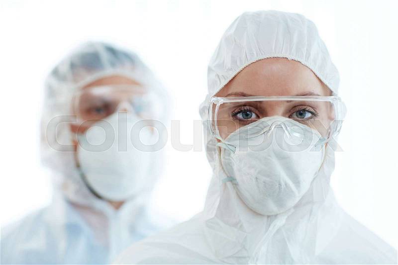 Young female in coveralls, eyeglasses and respirator looking at camera with her colleague on background, stock photo