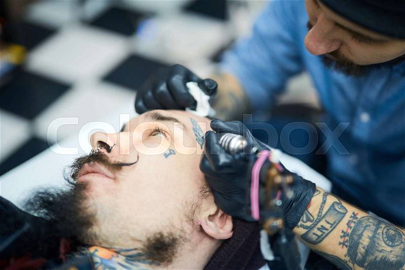 Master of tattoo art drawing on man face, stock photo