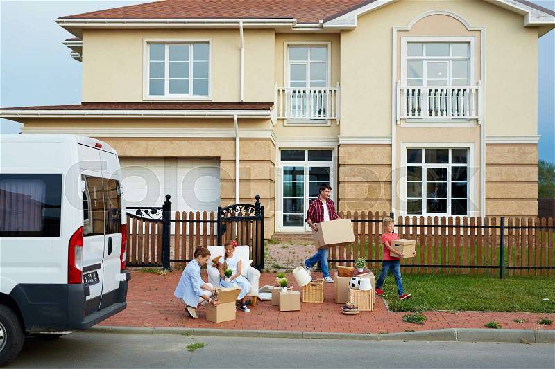 Portrait of family with two children moving out: mother and daughter busy sorting personal belongings in front of house while father and son taking out cardboard boxes to van outside, stock photo