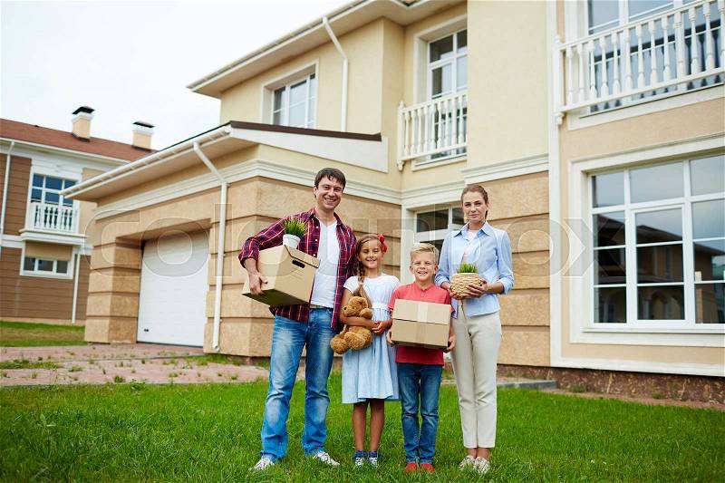 Portrait of successful family with two children standing together holding cardboard boxes on green grass lawn in front of their new house, smiling brightly looking at camera, ready to move in, stock photo