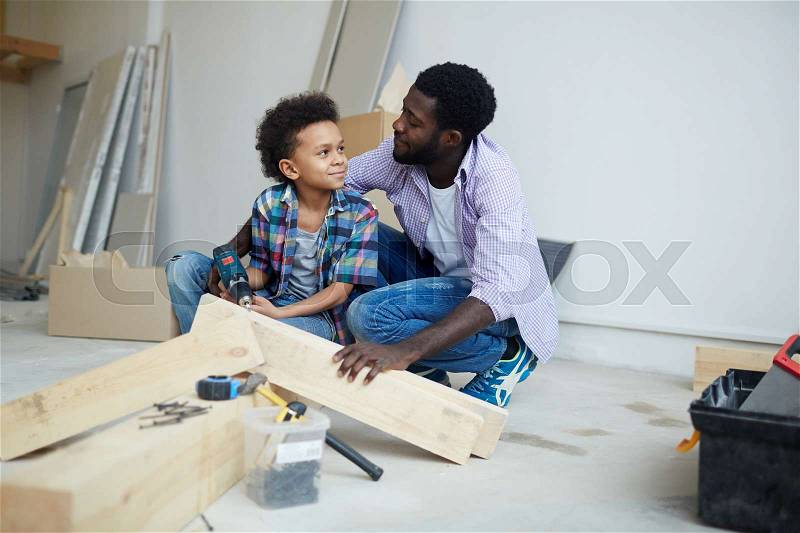 Cute boy talking to father while drilling wooden plank during house renovation, stock photo