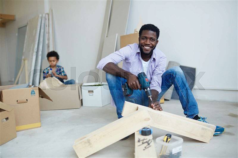 Young man drilling wooden planks while his son unpacking boxes on background, stock photo