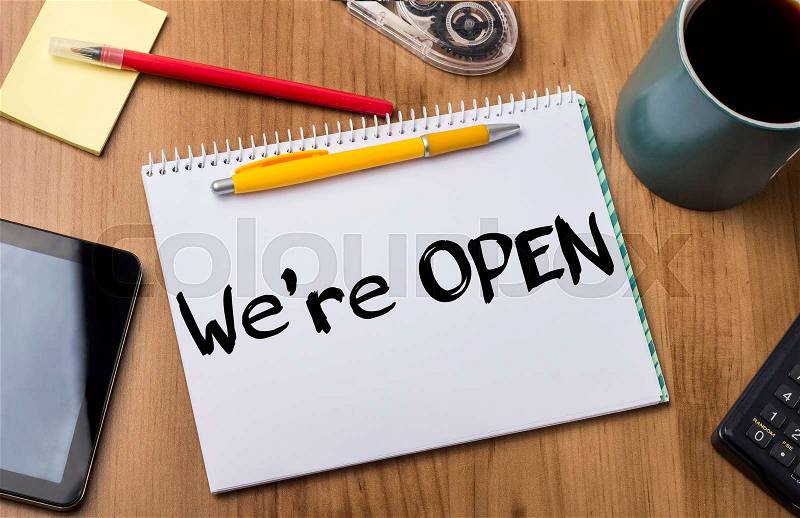 We’re OPEN - Note Pad With Text On Wooden Table - with office tools, stock photo