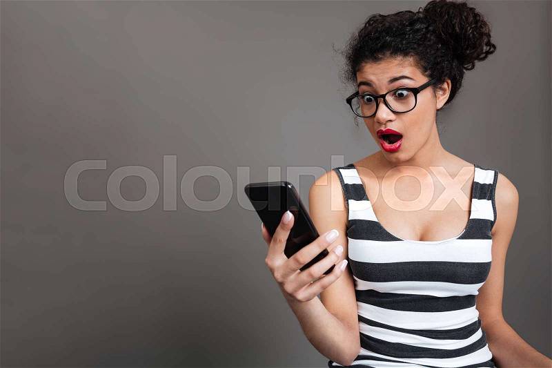 Portrait of a surprised african woman in eyeglasses looking at phone over gray background, stock photo
