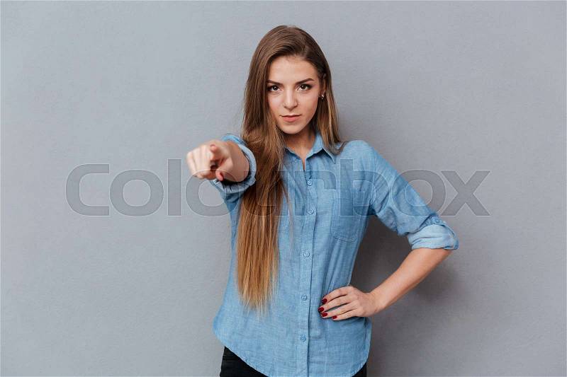Woman in shirt pointing at camera with arm at hip. Isolated gray background, stock photo