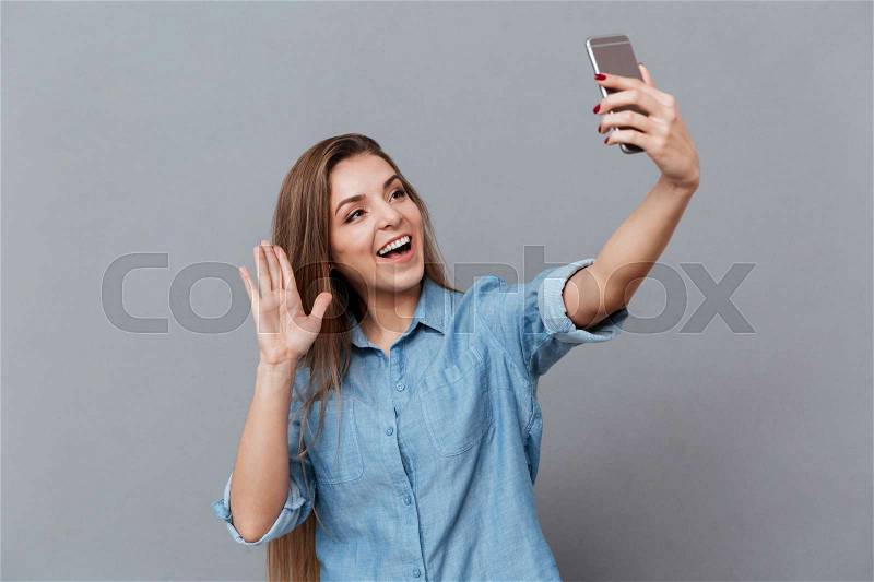 Happy Woman in shirt making selfie on smartphone and waving at camera. Isolated gray background, stock photo