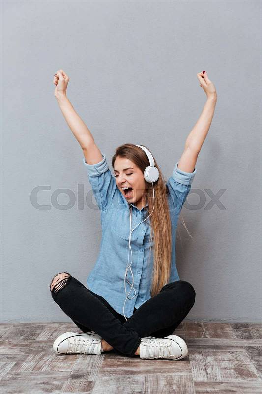 Vertical image of Woman in shirt listening to music on the wooden floor with hands up. Isolated gray background, stock photo