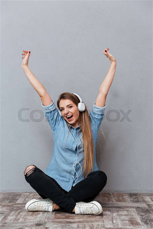 Vertical portrait of Happy Woman in shirt listening music and sitting on the wooden floor in studio. Isolated gray background, stock photo