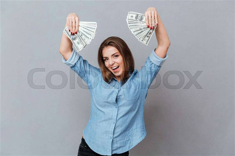 Happy Woman in shirt with money in hands and open mouth posing in studio. Isolated gray background, stock photo