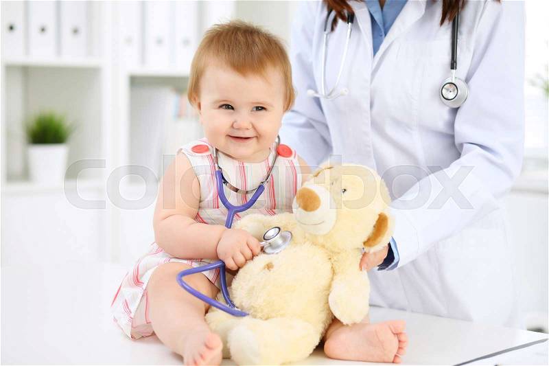 Happy cute baby at health exam at doctor's office. Toddler girl is sitting and keeping stethoscope and teddy bear , stock photo