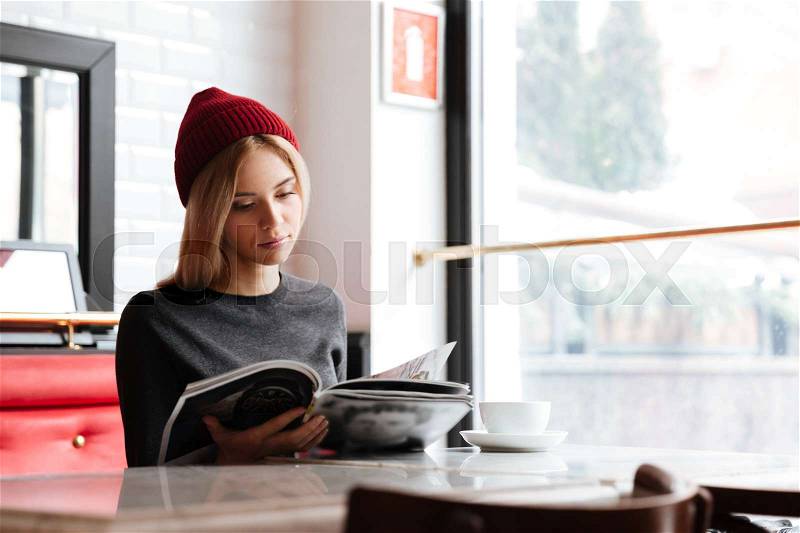 Young woman in red hat reading journal by the table in cafe near the window, stock photo