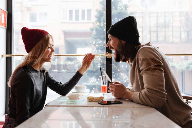 Side view of couple sitting by the table in cafe near the window. Woman feeding her boyfriend, stock photo