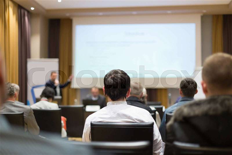 Presentation in lecture hall. Speeker having talk at public event. Participants listening to lecture. Rear view, focus on man in audience, stock photo