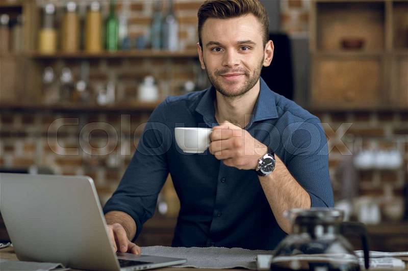Handsome young man with coffee cup using laptop and smiling at camera, stock photo