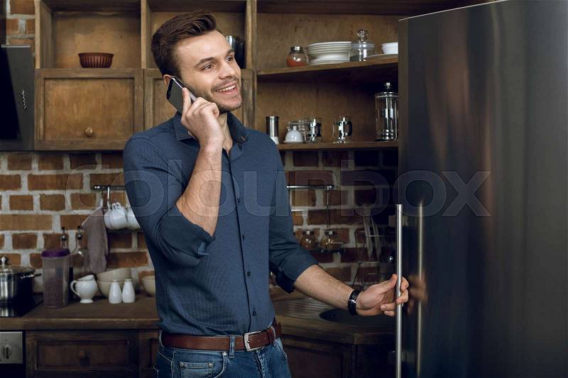 Happy young man talking on smartphone and looking away in kitchen, stock photo