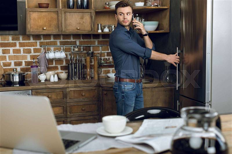 Pensive young man talking on smartphone while opening refrigerator in kitchen, stock photo