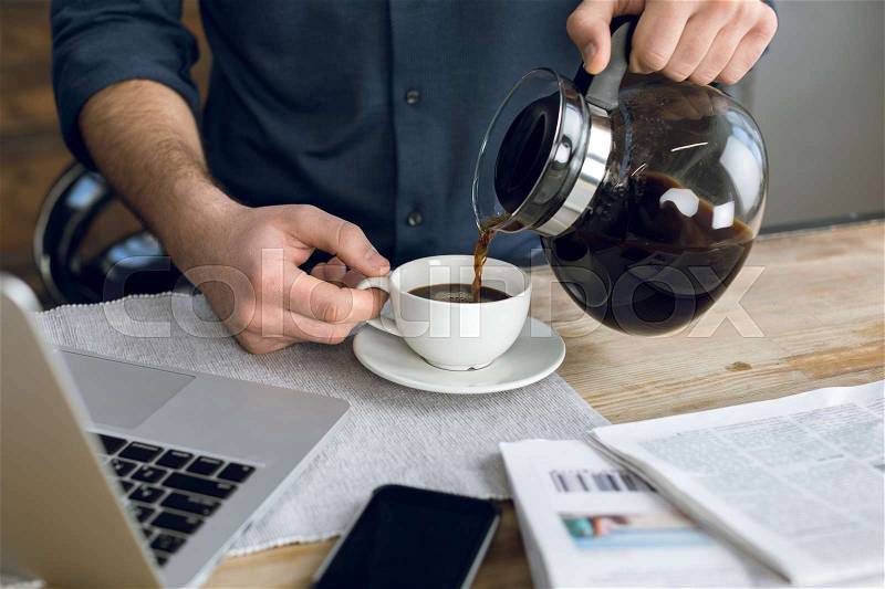 Partial view of man pouring coffee in cup on desk with laptop and smartphone, stock photo