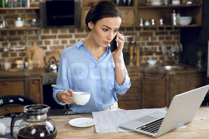 Young businesswoman with coffee cup using laptop and talking on smartphone, stock photo