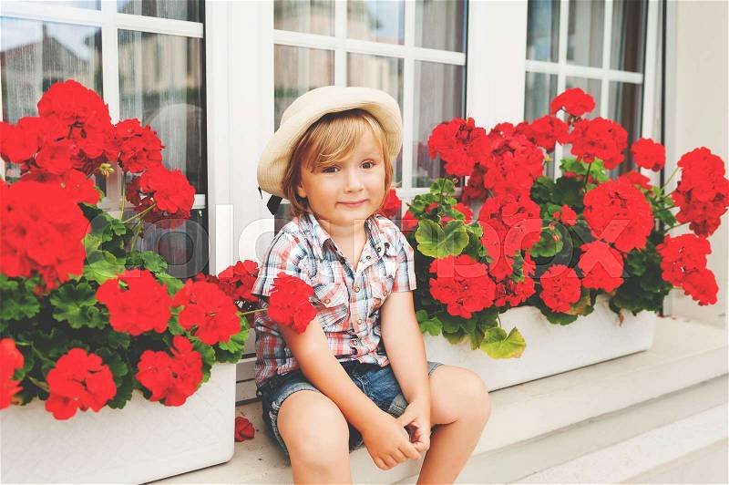 Adorable little boy gardener sitting between bright red geranium in pots, stock photo