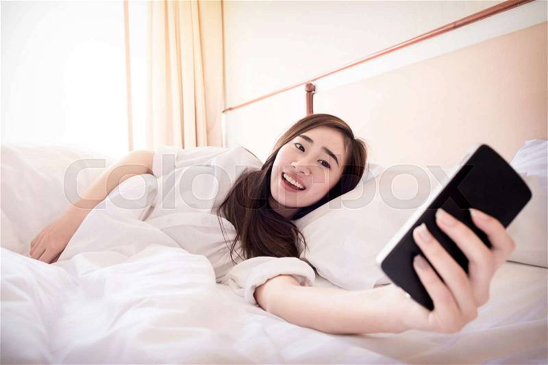 Relaxed woman at home reading a text message on smartphone in her bright bedroom, stock photo