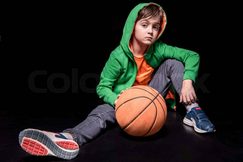 Pensive boy sitting on floor with basketball ball and looking to camera on black, stock photo