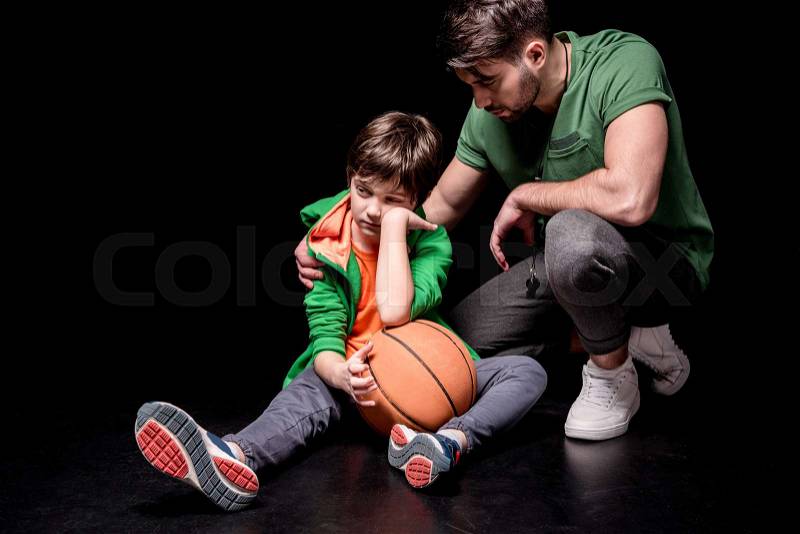 Man cheering up upset boy sitting on floor with basketball ball on black, stock photo