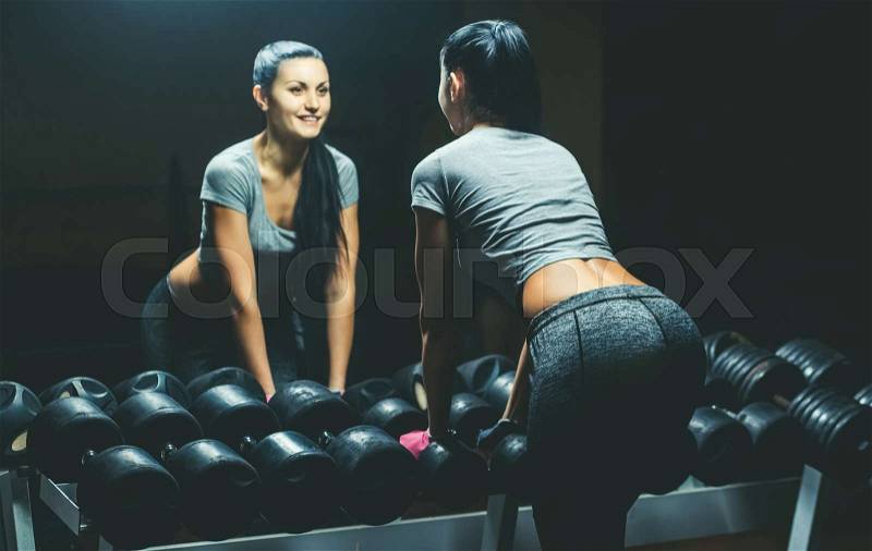 Slim, bodybuilder girl, lifts heavy dumbbell standing in front of the mirror while training in the gym. Sports concept, fat burning and a healthy lifestyle, stock photo