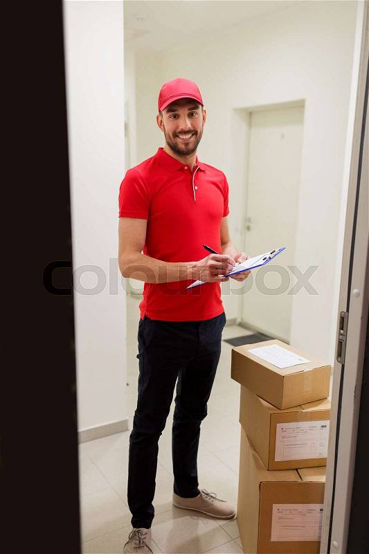 Delivery, mail, people and shipment concept - happy man in red uniform with parcel boxes and clipboard in corridor at open customer door, stock photo