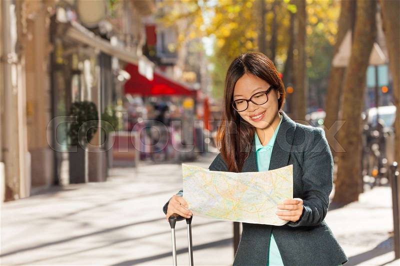 Pretty young Asian woman reading map walking through Paris with her suitcase, stock photo