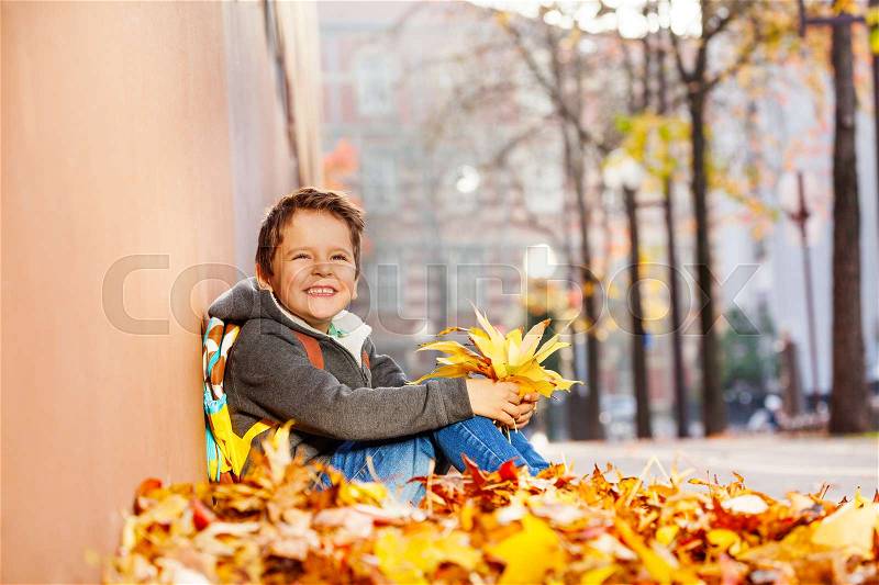 Smiling boy with rucksack sitting in leaf pile and holding autumn maple bouquet, stock photo